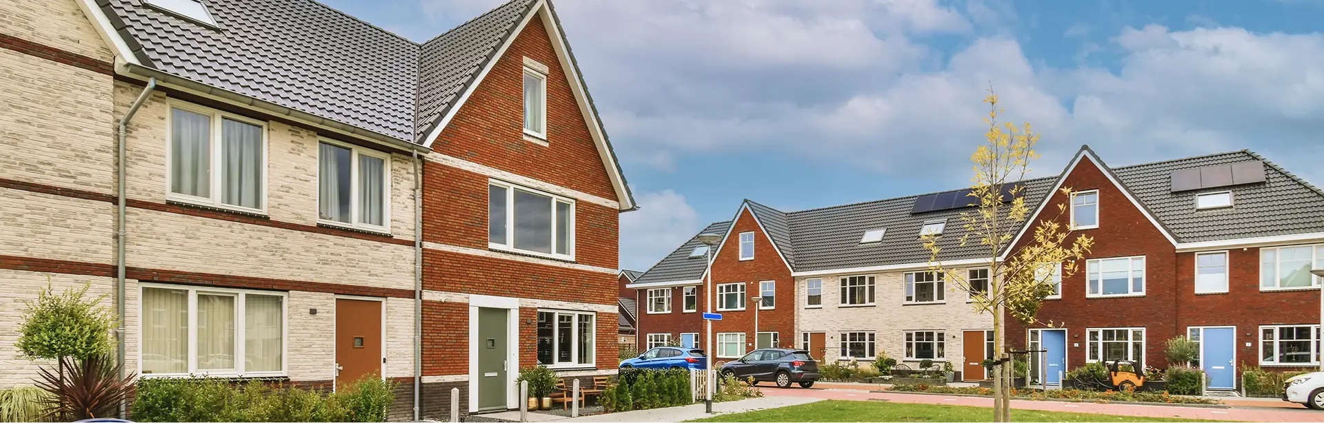 Modern brick-and-stone residential neighborhood with connected townhouses, green lawns, young trees, parked cars, and a partly cloudy blue sky.